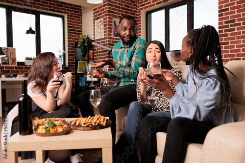 Culturally diverse friends talking, conversing each other, drinking wine, eating, tasting cheese, bread sticks at diverse people gathering. Different nationalities mates enjoying social life.