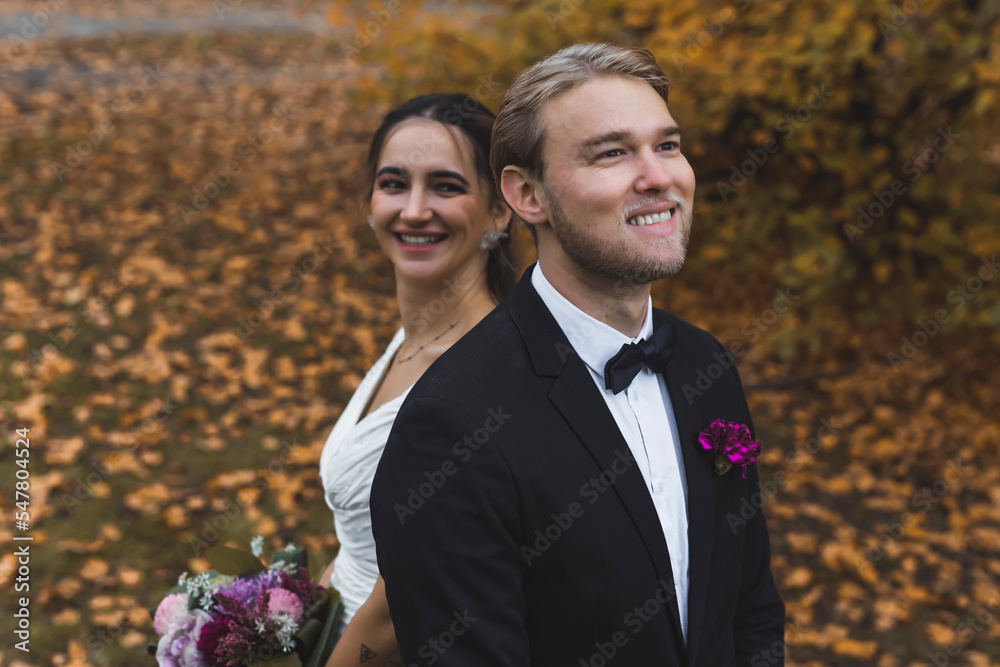 Happiest moment of life - wedding day with best friend. Dreamy blond bearded caucasian man in black tuxedo with butterfly tie standing in front of his smiling Turkish bride. Golden leaves on the grass Happiest moment of life - wedding day with best friend. Dreamy blond bearded caucasian man in black tuxedo with butterfly tie standing in front of his smiling Turkish bride. Golden leaves on the grass