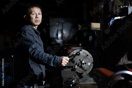 A craftsman poses in gray work clothes in front of a lathe at a local factory. Conceptual images of the essence of manufacturing, technical succession, and the challenge of high-precision machining.