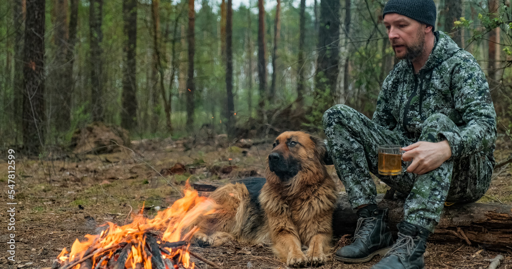 Adult Man is relaxing at the fire with his dog in the nature. Alone traveler is resting with his German shepherd by the forest fire and drinking tea. Man strokes a dog at the fire in the forest. Adult Man is relaxing at the fire with his dog in the nature. Alone traveler is resting with his German shepherd by the forest fire and drinking tea. Man strokes a dog at the fire in the forest.
