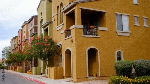 A street view of a row of warm colorful townhouses with stucco coating on the outside  arched doorways  and balconies with colorful red flowering trees in front.