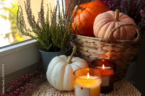 Beautiful heather flowers, burning candles and wicker basket with pumpkins near window indoors photo