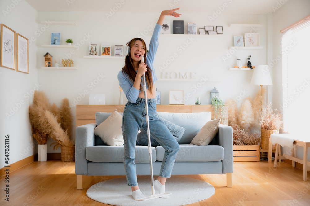 House cleaning with fun. Happy young asian housewife singing her favorite song during cleanup, using mop as microphone, enjoying domestic work. Young woman dancing and cleaning in living room - obrazy, fototapety, plakaty 