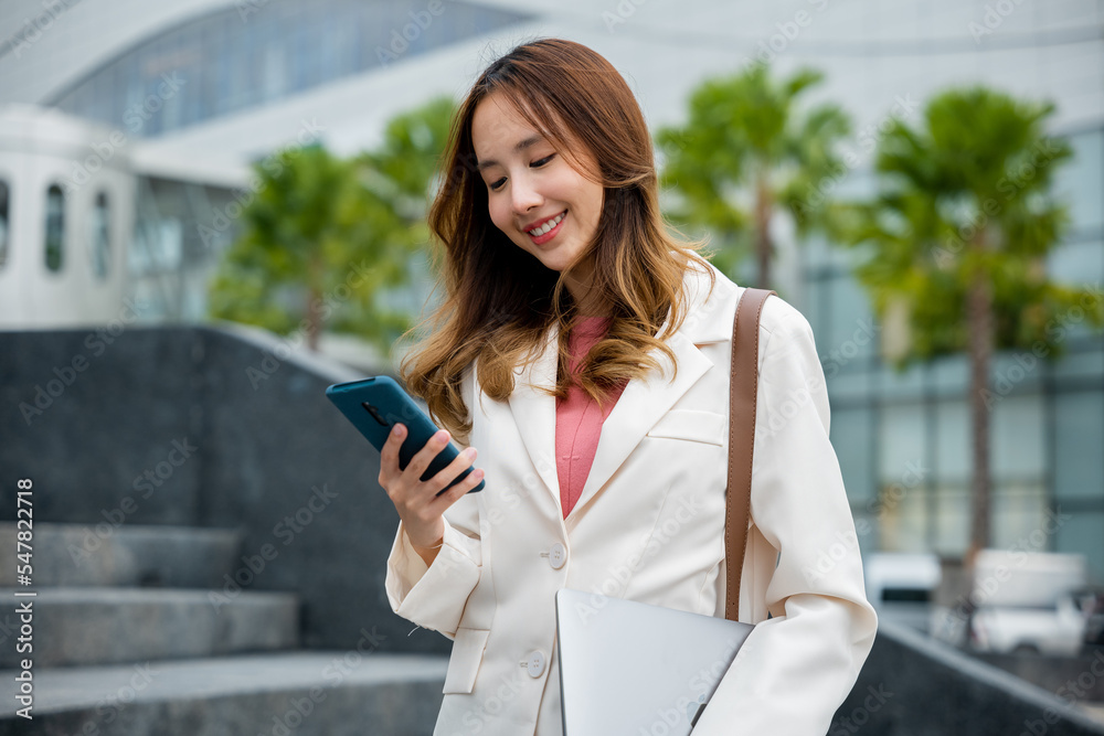 Asian woman successfully work go to against city scene of modern office buildings in the city. Portrait of confidence Asian businesswoman holding laptop and check message on mobile smart phone. Asian woman successfully work go to against city scene of modern office buildings in the city. Portrait of confidence Asian businesswoman holding laptop and check message on mobile smart phone.