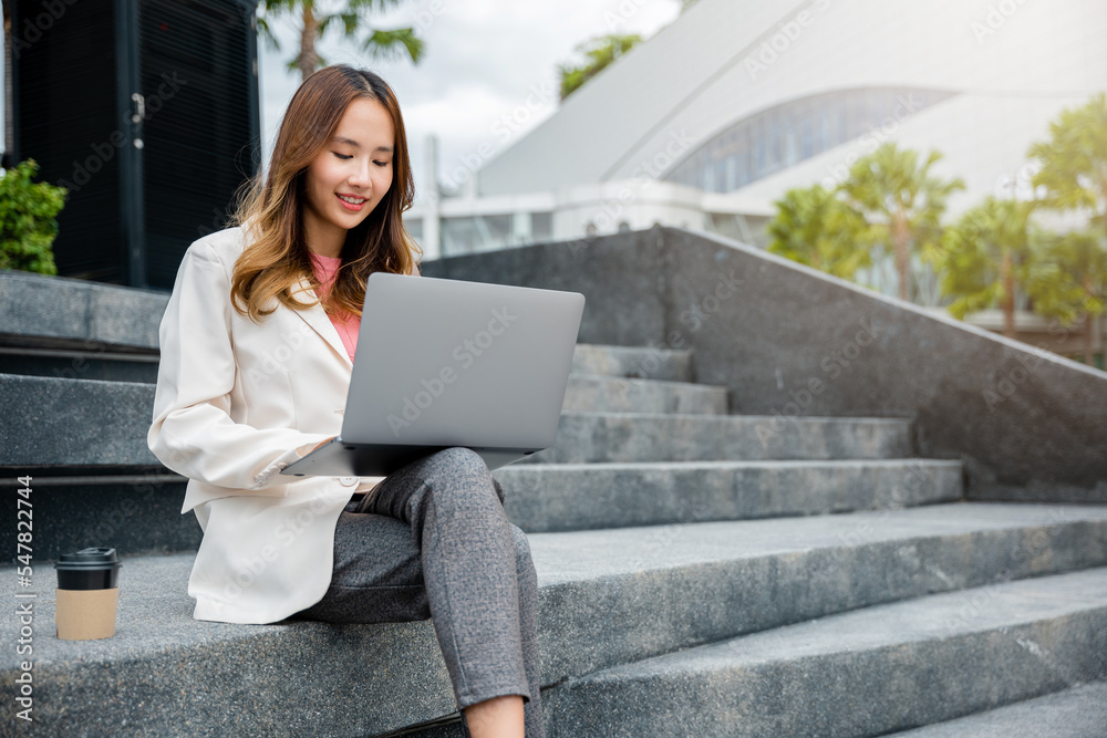 Remote working concept outside the office. Happy young Asian woman freelance smile looking laptop computer with smile while working with coffee cup while sitting on stairs outdoors in the city. Remote working concept outside the office. Happy young Asian woman freelance smile looking laptop computer with smile while working with coffee cup while sitting on stairs outdoors in the city.