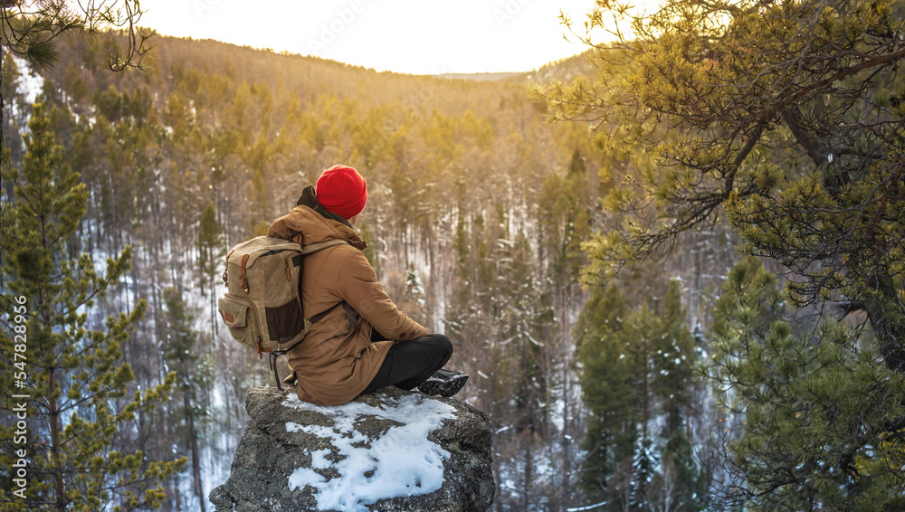 Fototapeta premium A tourist-traveler with a backpack in a red hat sits on the edge of a cliff and looks at a forest winter valley