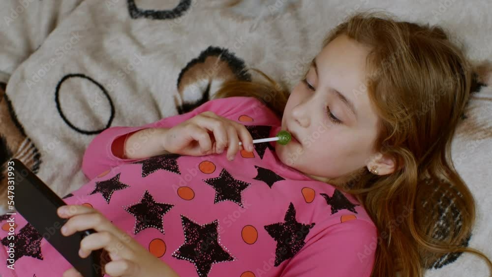 A cute girl with long hair is lying on the sofa, carefully looking at the smartphone screen and sucking on a sweet lollipop. The concept of digital addiction in children and adolescents