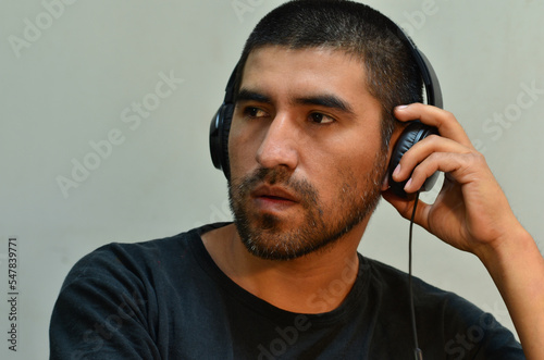 Hombre latino joven con pelo corto y auriculares escuchando música con fondo blanco y remera negra 