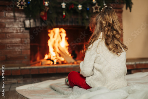 A happy little girl is sitting by the fireplace in a cozy living room on Christmas Eve. The concept of Christmas and New Year. Rear view