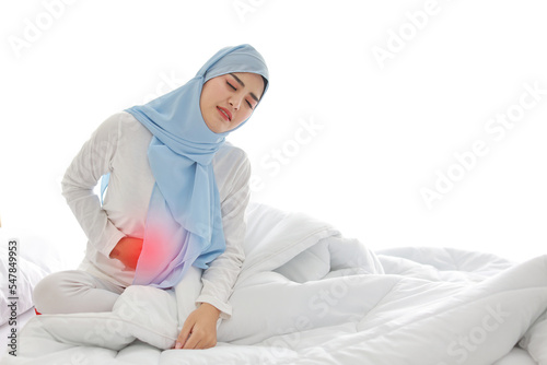 Menstrual pain or stomachache young asian muslim woman sitting on bed. Beautiful girl hold stomach and feeling painful with unhappy face. Healthy concept photo