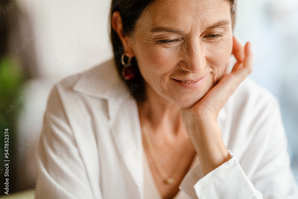 White mature woman in shirt smiling while sitting in cafe indoors White mature woman in shirt smiling while sitting in cafe indoors