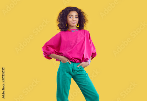 Studio portrait of beautiful stylish dark-skinned curly preteen girl isolated on yellow background. Ethnic girl in fashionable colored teenage clothes and with accessories smiling at camera.