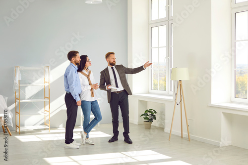 Photographie Real estate agent young man in suit showing new big modern flat to couple of buyers or tenants