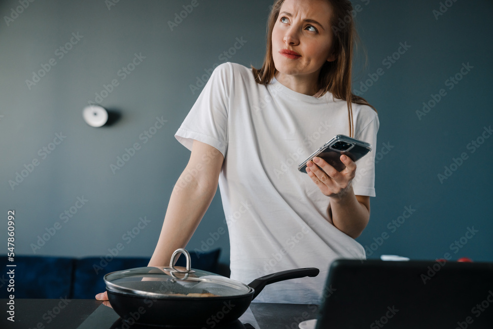 White puzzled woman using cellphone and cooking breakfast White puzzled woman using cellphone and cooking breakfast