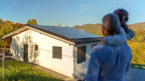 Rear view of dad holding her little girl in arms and looking at their house with installed solar panels. Alternative energy, saving resources and sustainable lifestyle concept.