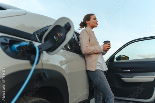 Young woman with cup of coffee waiting while her electric car charging, sustainable and economic transportation concept.