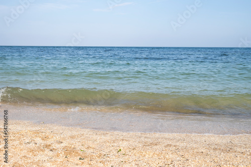 Birose blue sea, golden coast of small shells and sand, a clear horizon line and a clear sky. Soft sunlight, clear sky with glowing clouds, waves and water splashes