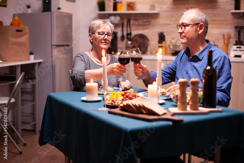 Elderly couple dining together with wine in kitchen. Happy cheerful senior elderly couple dining together in the cozy kitchen  enjoying the meal  celebrating their anniversary.