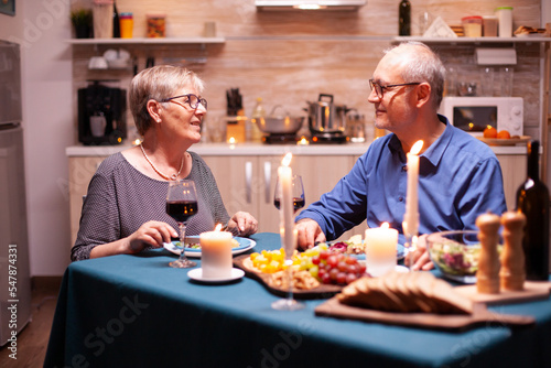 Having a conversation during din romantic dinner. Happy cheerful senior elderly couple dining together in the cozy kitchen, enjoying the meal, celebrating their anniversary.