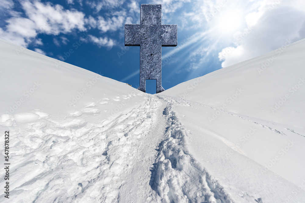 Large stone religious cross with a door and a trail with footprints in the snow in a mountain landscape against a blue sky with clouds, sun rays and copy space. Large stone religious cross with a door and a trail with footprints in the snow in a mountain landscape against a blue sky with clouds, sun rays and copy space.