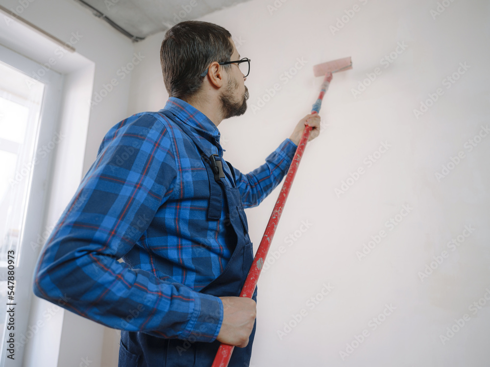 young man in blue work suit hold paint roller, over white wall room background. Instruments accessories for renovation apartment room. Repair home concept young man in blue work suit hold paint roller, over white wall room background. Instruments accessories for renovation apartment room. Repair home concept