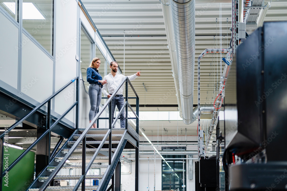 Business colleagues reviewing machinery standing on staircase in factory Business colleagues reviewing machinery standing on staircase in factory