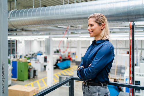 Smiling businesswoman standing with arms crossed near railing in factory