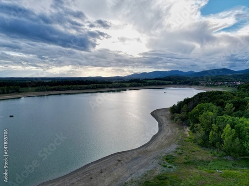 Flight over the Michelbach lake dam (Upper-Rhine, Alsace, France) by drought and cloudy weather in summer with a very low water level, in the evening at sunset with village and mountains in background