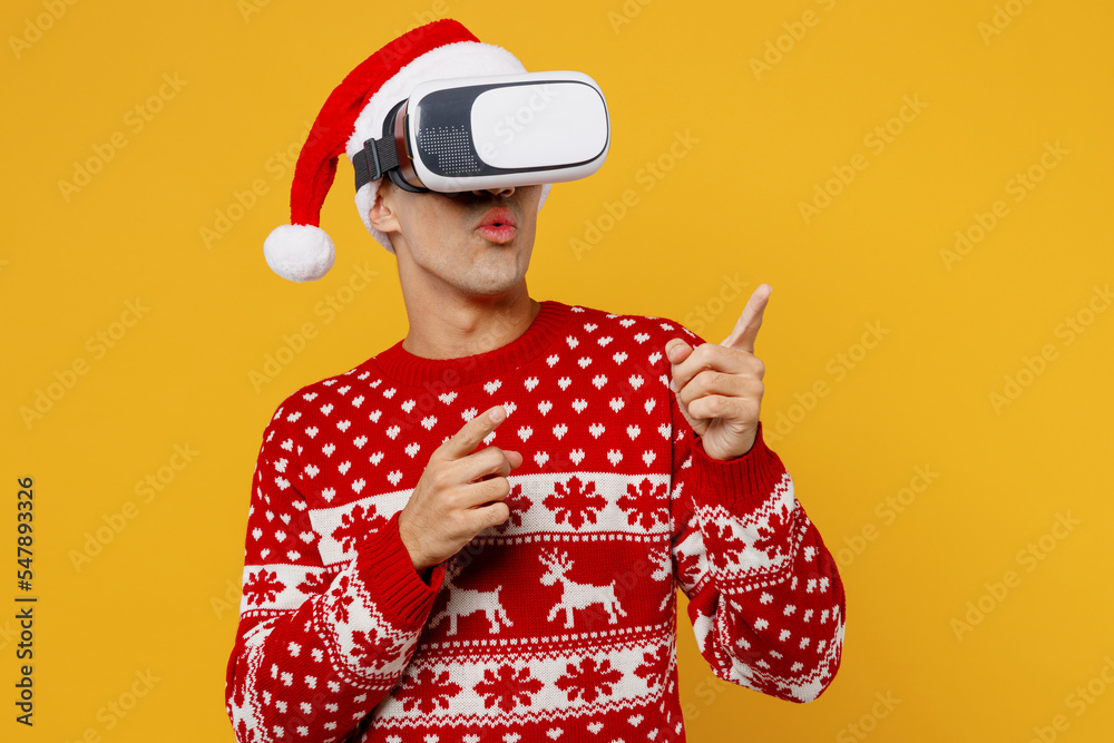 Merry young man wear red knitted christmas sweater Santa hat posing watching in vr headset pc gadget point finger aside on area isolated on plain yellow background Happy New Year 2023 holiday concept. Merry young man wear red knitted christmas sweater Santa hat posing watching in vr headset pc gadget point finger aside on area isolated on plain yellow background Happy New Year 2023 holiday concept.
