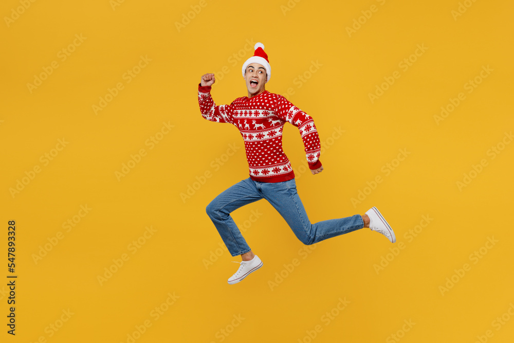 Full body side view merry young man wear red knitted Christmas sweater Santa hat posing run look back aside on area isolated on plain yellow background Happy New Year 2023 celebration holiday concept Full body side view merry young man wear red knitted Christmas sweater Santa hat posing run look back aside on area isolated on plain yellow background Happy New Year 2023 celebration holiday concept