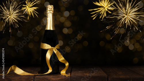 New Year Sylvester celebration holiday New Year's Eve greeting card - Champagne or sparkling wine bottle with golden gift ribbon on old rustic wooden table and pyrotechnics firework in the background