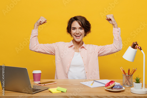 Young successful employee business woman wear casual shirt sit work at office desk with pc laptop showing biceps muscles on hand isolated on plain yellow color background. Achievement career concept.
