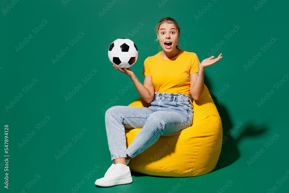 Full body young woman fan wear basic yellow t-shirt cheer up support football sport team sit in bag chair hold soccer ball watch tv live stream spread hands isolated on dark green background studio. Full body young woman fan wear basic yellow t-shirt cheer up support football sport team sit in bag chair hold soccer ball watch tv live stream spread hands isolated on dark green background studio.