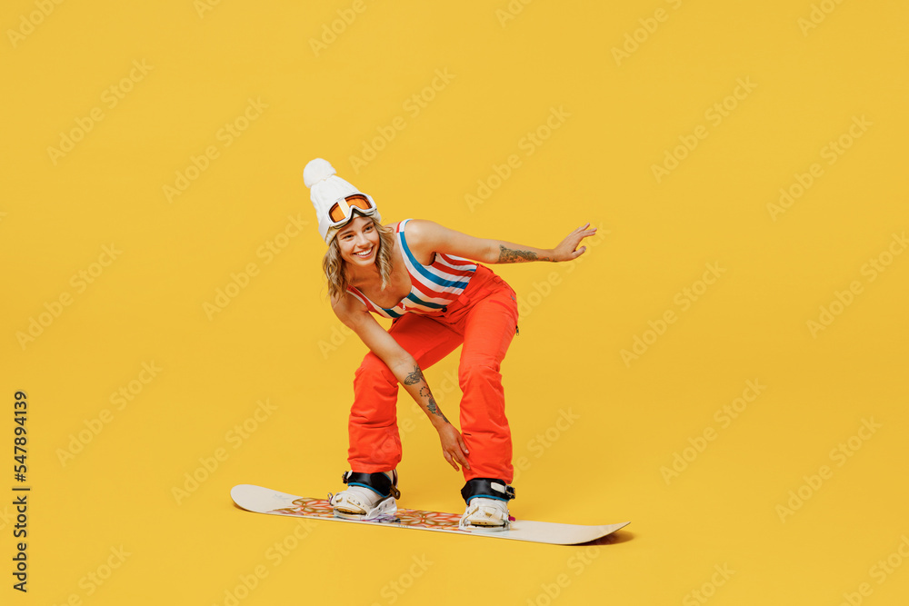 Snowboarder fun happy woman wear orange ski suit goggles mask hat swimsuit spend extreme weekend snowboarding leaning isolated on plain yellow background studio. Winter sport hobby trip relax concept.