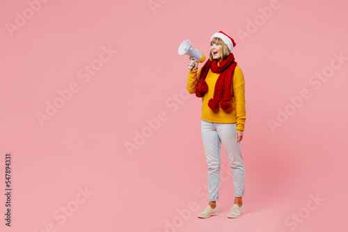 Full body elderly woman 50s year old wear knitted sweater red scarf Santa hat posing hold scream in megaphone announces sale isolated on plain pink background. Happy New Year Christmas 2023 concept.