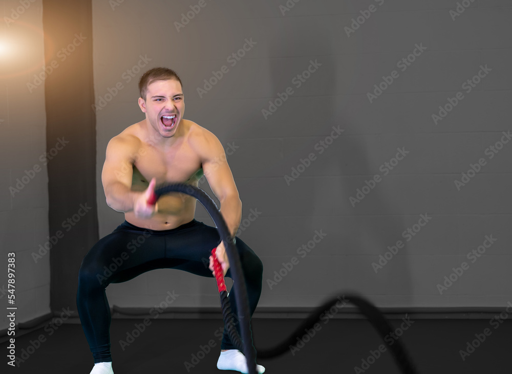 A handsome muscular young man using battle ropes for exercise in a gym. He focuses on performing the exercise, screams. Motivational photo. Copy space. A handsome muscular young man using battle ropes for exercise in a gym. He focuses on performing the exercise, screams. Motivational photo. Copy space.