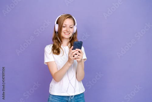 Young woman watching video clips on the phone in wired headphones in casual clothes with a pensive look on a colored background in the studio
