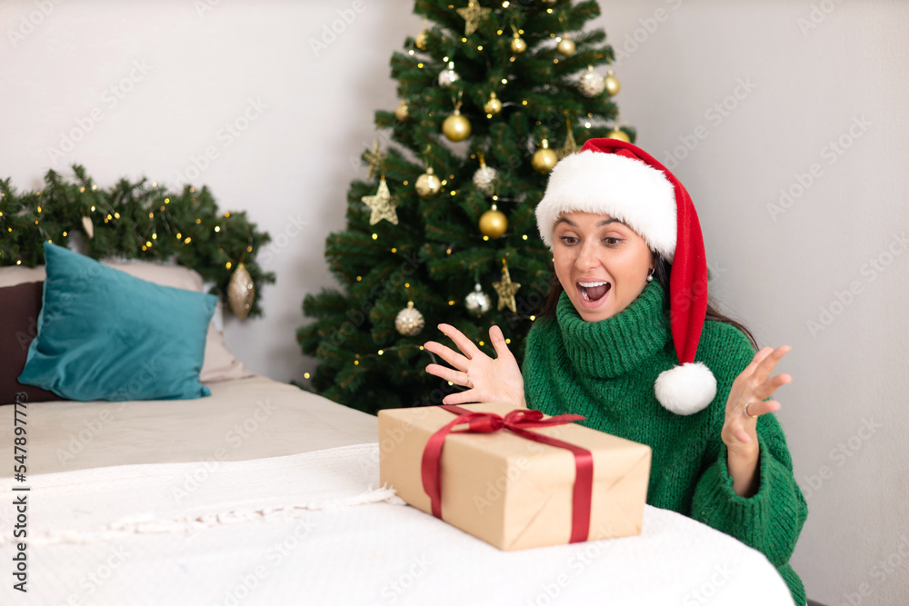 Merry Christmas. Portrait of a beautiful young woman in a cozy bedroom with a decorated Christmas tree in the background. Green sweater. Lifestyle