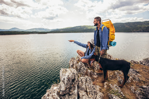 Hikers with backpacks and their doberman sitting on cliff enjoying at the mountain lake