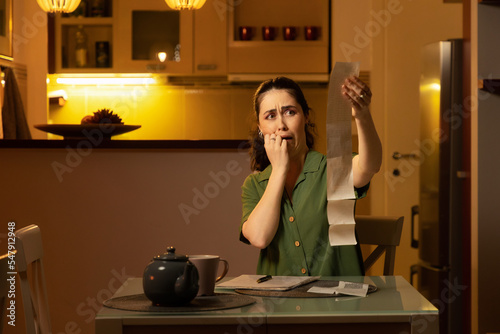 Utiliti bills. Frightened young Caucasian woman is sitting at table and checking expenses looks on receipt. Concept of accounting and household expenses photo