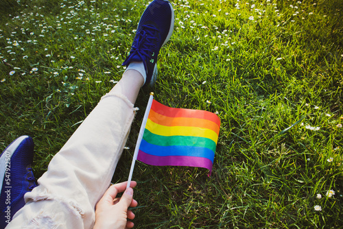 LGBT flag in rainbow colors in female hand top view. A lesbian, bisexual, gay girl sits relaxing in green clearing in park on sunny day. Pride Month in June. Faceless woman on picnic. Legs in jeans.
