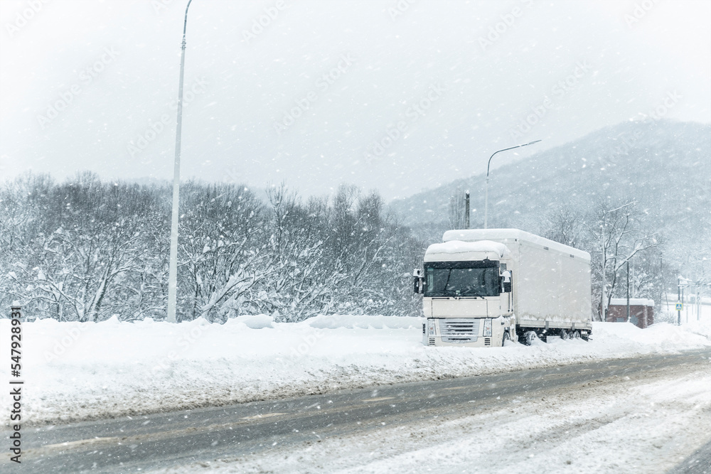 Obraz premium Big commercial semi-trailer truck trapped in snow drift on closed highway road at heavy snow storm blizzard cold winter day. Cargo vehicle stuck on freeway at bad weather conditions frosty snowfall