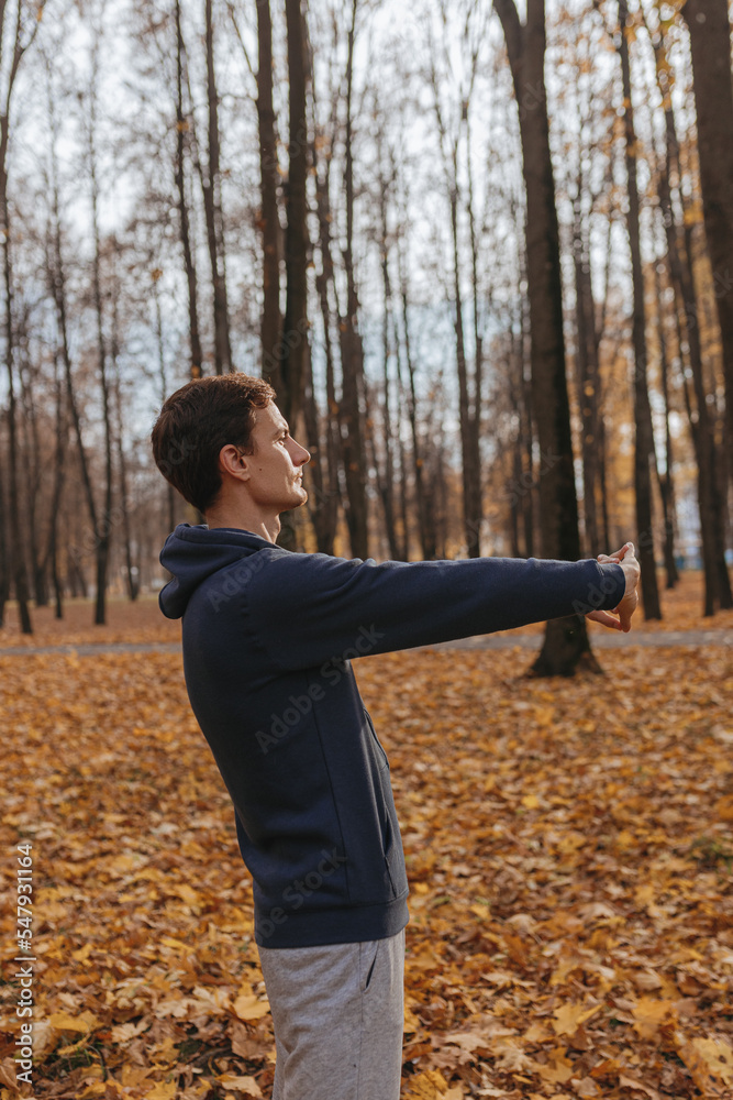 Side view of active sportsman stretching arms and warming up during training on sunny day in autumn park Side view of active sportsman stretching arms and warming up during training on sunny day in autumn park