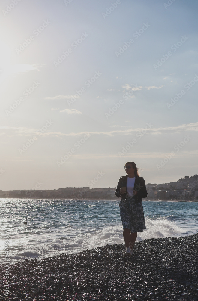 Young beautiful girl with coffee on an autumn sunny day walks on the beach of the Mediterranean Sea