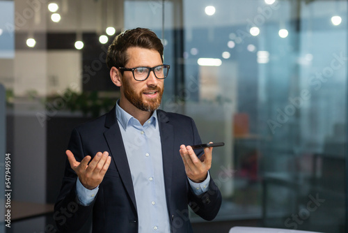 Foto Friendly and smiling businessman in office recording audio message, man using smartphone app for communication, investor in business suit near window inside building