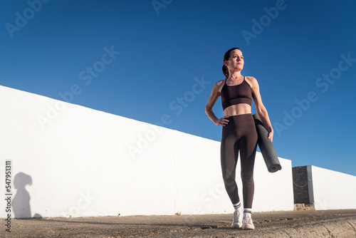 Fit sporty woman carrying a rolled up exercise mat outside.