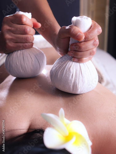 A traditional Thai massage with hands of therapist holding herbal hot compress balls on a back of woman for induces deep relaxation, relieves stress and fatigue in spa salon