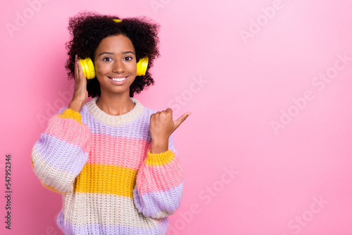 Portrait of cheerful girl with wavy hairdo wear knit sweatshirt touch headphones directing empty space isolated on pink color background