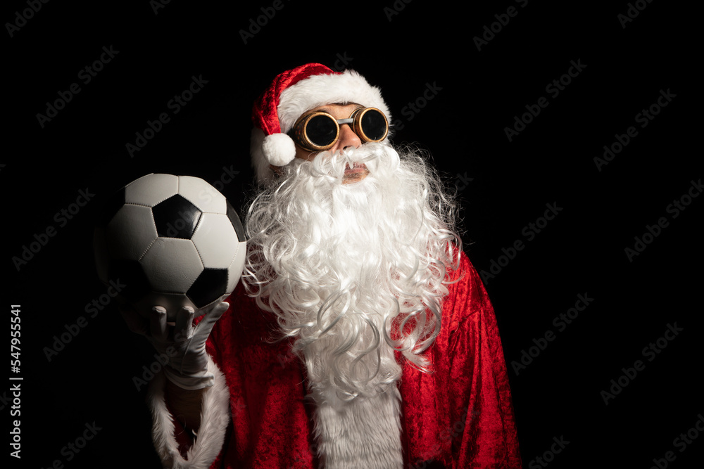 Santa Claus with sunglasses or a welder holding a World Cup soccer ball as a gift for all children, with a black background where his red suit and white beard stand out Santa Claus with sunglasses or a welder holding a World Cup soccer ball as a gift for all children, with a black background where his red suit and white beard stand out