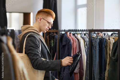 Side view portrait of young man looking at clothes while shopping sustainably in thrift store photo